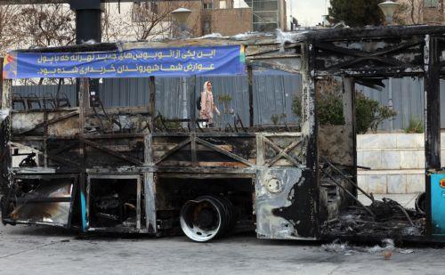 epa12650622 An Iranian woman walks near the wreckage of a burnt-out public bus in Tehran, Iran, 15 January 2026. The country remains under a near-total nationwide internet blackout that began on 08 January, amid an intensifying wave of anti-government protests.  EPA/ABEDIN TAHERKENAREH